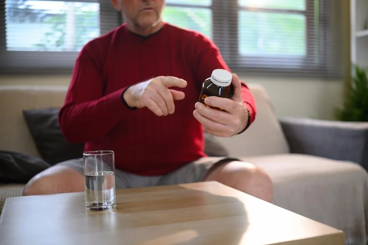 Dojrzały mężczyzna przyglądający się butelce z receptą przy stole w domu z szklanką wody
